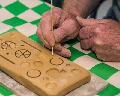 Een persoon tekent patronen op een blok klei met een houten stokje, terwijl ze op een groen-wit geruit tafelblad zitten.