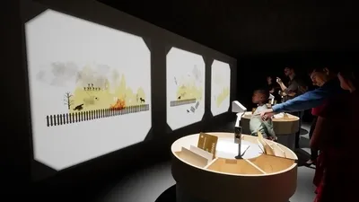 Interactieve tentoonstelling met projecties en aanwezigen die deelnemen aan activiteiten rond een centrale tafel.