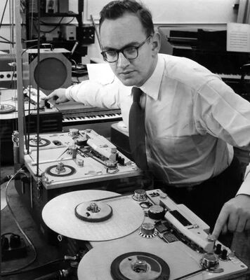 Man in een kantoor, bezig met een analoge tape recorder en andere audioapparatuur. Achtergrond met een toetsenbord zichtbaar.