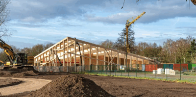 Nieuwbouw van een houten constructie met een kraan en bouwmachines op een bouwplaats, omgeven door een groene omgeving.